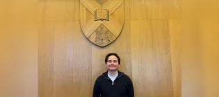 A male student standing against the University of Edinburgh logo on the wall